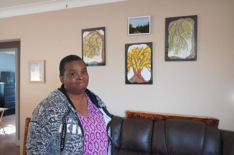 an African American woman wearing a lilac colored shirt and black and white zip-up hoodie stands in front of a beige wall where four artworks she made hang. She smirks at the camera as she poses stoically in front of the art.
