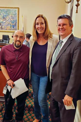 Three people stand next to each other, smiling at the camera. On the far left, a man wearing a burgundy polo and glasses holds a cane and papers in one hand. On the far right, a man wearing a gray suit and blue striped tie holds a paper in one hand. In the middle, a woman wearing a navy t-shirt and khaki blazer has her arms on either side of the men.