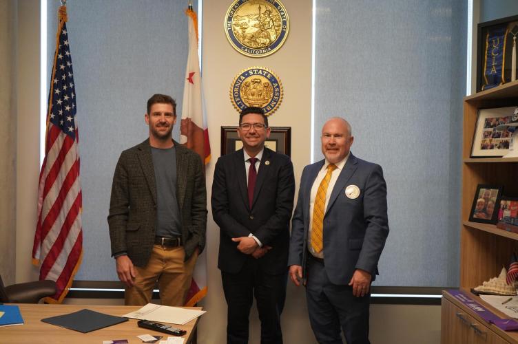 Three men wearing suits stand next to each, smiling at the camera. They stand in a legislative office in front of two windows on the left and right, an American flag on the left, and a bookshelf on the right. In the center behind the men, two gold California seals trimmed with blue are hung on the wall. 