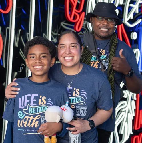 a family with beaming smiles wearing matching navy t-shirts with text reading life is better with best buddies pose in front of a red, white, blue, and black background
